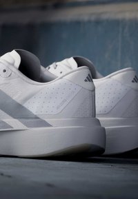 Ζευγάρι λευκά παπούτσια τρεξίματος Adidas Adizero Evo SL, που φαίνονται από πίσω πάνω σε γκρι επιφάνεια με θολό σκοτεινό φόντο.