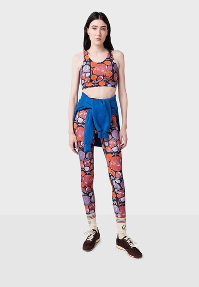 Soutien-gorge de sport à motif floral et leggings de couleur violet foncé, orange et rose, avec un pull bleu noué à la taille. Chaussettes blanches à rayures et baskets.