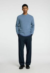 Helles blaues Strickpullover mit runder Ausschnitt, kombiniert mit dunkelblauen Hosen. Schwarze Schuhe mit strukturiertem Detail. Schlichtes, modernes Design.