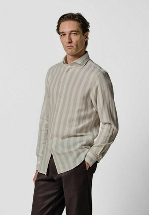 Hellbeiges Hemd mit Knöpfen, vertikalen, dezenten Streifen; lange Ärmel und spitzer Kragen; kombiniert mit dunkelbraunen Hosen.