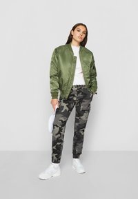 Olivgrüne Bomberjacke mit Satin-Finish und Reißverschluss. Camouflage-Cargohose mit elastischen Bündchen. Weiße Sneakers. Einfaches weißes Oberteil unter der Jacke.