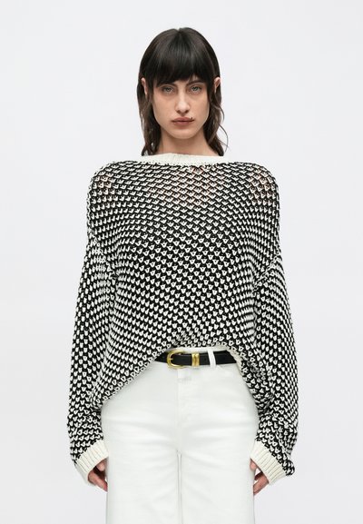 Pull oversize tricoté en noir et blanc avec une coupe ample, présentant un motif distinctif, un col côtelé et des manches larges. Associé à un pantalon blanc.