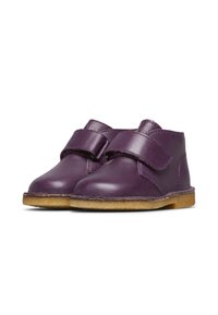 Un paio di scarpe in pelle viola per bambini con fasce in Velcro e suole in crepe marrone chiaro su uno sfondo bianco.