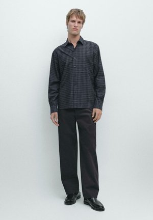 Jonge man staand tegen een witte muur, draagt een donker geruit overhemd, zwarte wijde broek en zwarte schoenen, met een neutrale uitdrukking.