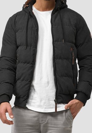 Mand iført en sort vatteret jakke med lynlås og hætte over en hvid t-shirt og grå bukser, den ene hånd i lommen.