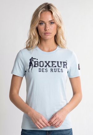 Blonde woman wearing light blue t-shirt with "Boxeur des Rues" text and a boxing figure graphic, standing against a plain background.