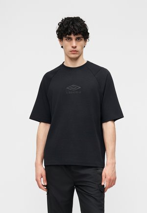 Νεαρός άνδρας με σγουρά μαλλιά, ντυμένος με μαύρο κοντομάνικο πουκάμισο Umbro και μαύρο παντελόνι, στέκεται μπροστά σε ένα απλό ανοιχτό φόντο.