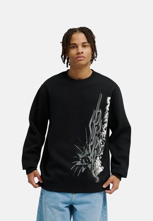 Person mit kurzen Twists trägt ein schwarzes Sweatshirt mit weißen Flammengrafiken und Text, dazu eine hellblaue Jeans, stehend vor einem schlichten Hintergrund.