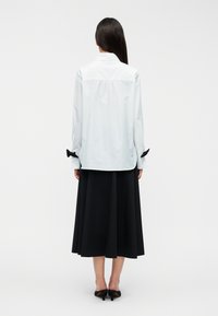 Φωτεινό μπλε πουκάμισο με γιακά και κούμπωμα κουμπιών στο πίσω μέρος, με μαύρες διακοσμητικές φιόγκους στα μανικέτια. Συνδυάζεται με μαύρη φούστα πλήρους μήκους.