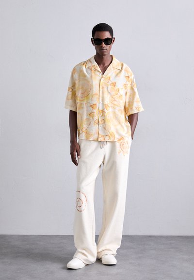 Bărbat îmbrăcat cu ochelari de soare negri, o cămașă scurtă cu mâneci scurte, decolorată, cu un model galben, pantaloni largi crem cu desene portocalii spiralate și adidași albi.
