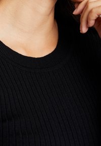 Gros plan sur une personne portant un pull noir côtelé avec un col rond, une main levée près du visage.