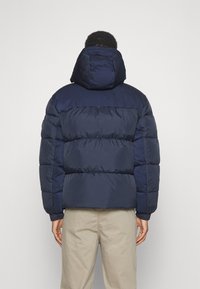 Marineblå puffjakke med polstret design og avtagbar hette, med elastiske mansjetter og en glatt finish. Brukt sammen med beige bukser.