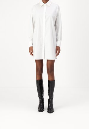 Femme portant une robe chemise blanche à manches longues avec col, associée à des bottes en cuir noires montantes jusqu'aux genoux, debout sur un fond blanc.