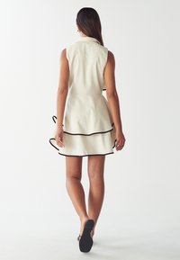 Donna che si allontana indossando un vestito senza maniche color crema con bordi neri e una gonna a strati, abbinato a tacchi aperti neri e orecchini a cerchio dorati.