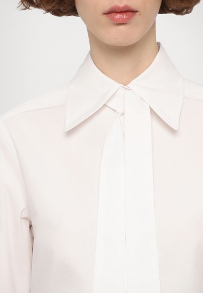 Blusa bianca con colletto strutturato e dettaglio a nodo al collo. Presenta un tessuto liscio e leggero e un design pulito e minimale.