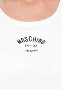 Valge soonikkoes tank top, millel on kaarjas dekoltee ja must trükitud tekst: "MOSCHINO 86 • 23 JEANS", mis asub dekoltee all keskel.