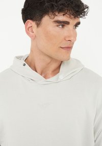Ljusgrå hoodie med en strukturerad yta, med en huva, tryckknappar och en broderad logotyp på bröstet.