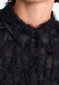 Sort bluse med et tekstureret design, der har delikate stoffringe og en traditionel krave, afsluttet med en sort knaplukning.