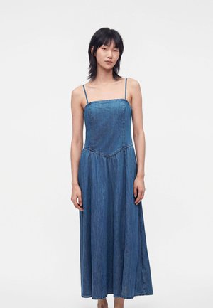 Person står mod en ensfarvet baggrund iført en ærmeløs, ankellang blå denimkjole med spaghetti-stropper og vender fremad.