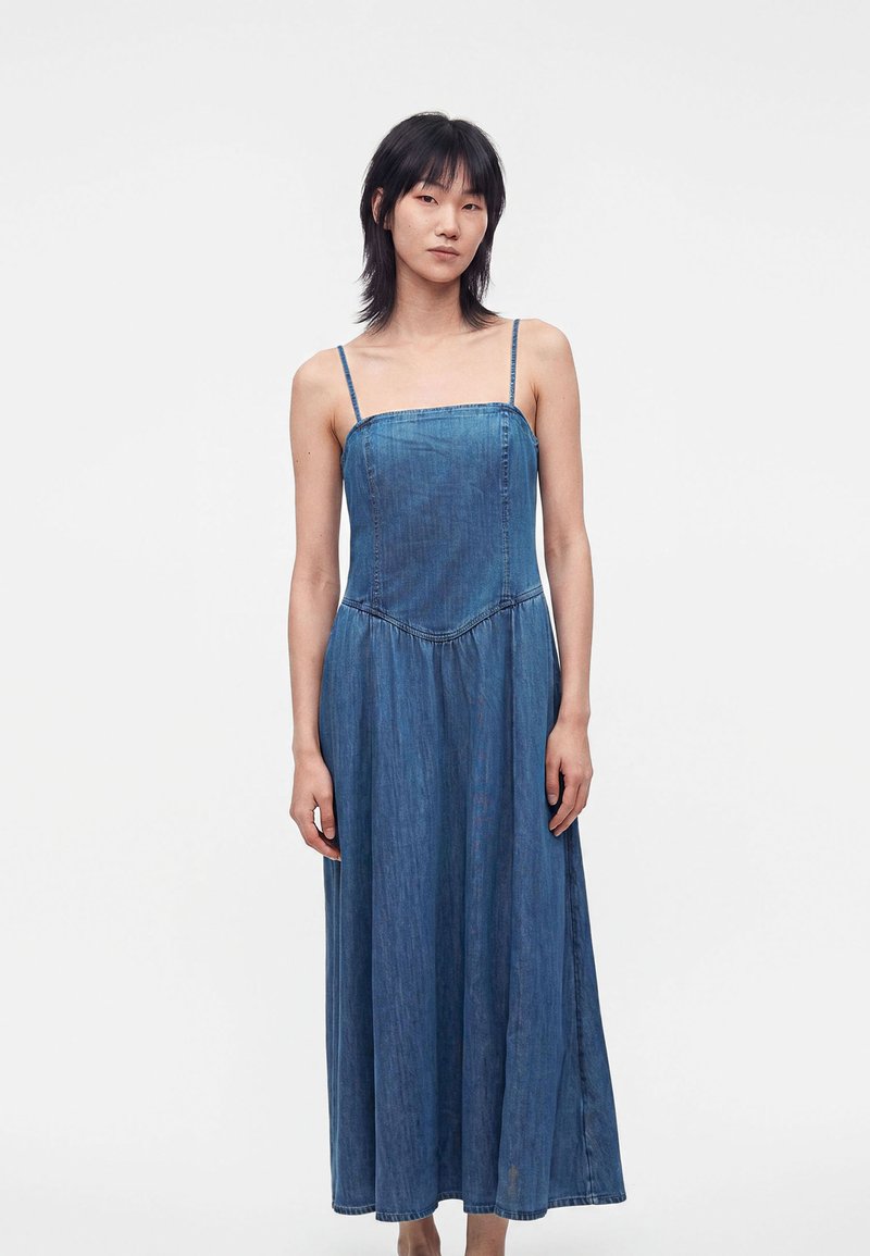 Personne debout devant un fond uni, portant une robe en jean bleu sans manches, longue jusqu’aux chevilles, avec des bretelles fines, tournée vers l’avant.