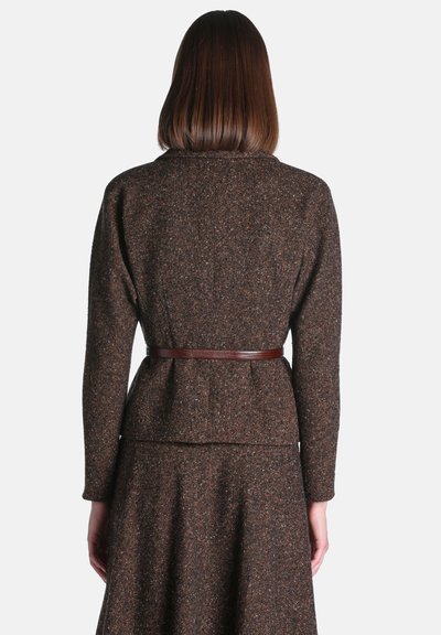 Femme vue de dos portant un blazer en tweed marron foncé et une jupe assortie avec une fine ceinture marron à la taille sur un fond uni.