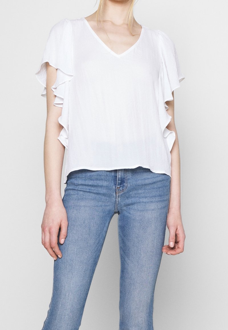 Femme portant un chemisier blanc à col en V avec des manches courtes à volants et un jean en denim bleu clair sur un fond uni.