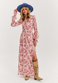 Dlouhé šaty s květinovým vzorem v růžových a oranžových tónech, s dlouhými rukávy a bočním rozparkem, doplněné širokým modrým kloboukem a béžovými botami.