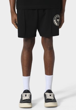 Persona che indossa pantaloncini neri con un logo circolare sulla gamba sinistra, calzini bianchi al polpaccio e sneaker basse nere con lacci e suole bianche.