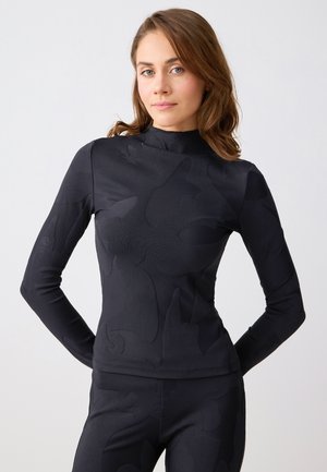 Kobieta o jasnobrązowych włosach, ubrana w dopasowaną czarną bluzkę z długim rękawem i pasujące spodnie z subtelnymi abstrakcyjnymi wzorami na neutralnym tle.