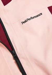 Giacca rosa Peak Performance con accenti bordeaux, chiusura con zip, texture liscia e dettaglio logo ricamato. La zip presenta accessori neri.