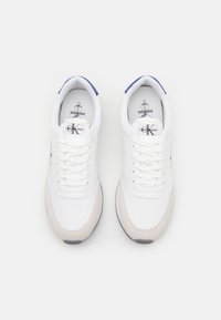 Zapatillas deportivas blancas con un acabado suave. Presentan una puntera de ante beis, acentos azules en el talón y un diseño con cordones.
