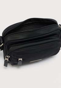 Μαύρη τσάντα ώμου από νάιλον με δύο θήκες με φερμουάρ, ασημένιες μεταλλικές λεπτομέρειες και λεία επιφάνεια. Συμπαγές ορθογώνιο σχήμα.