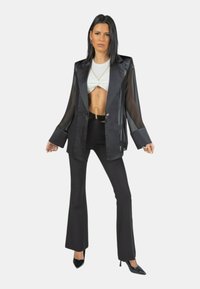 Blazer nero trasparente con revers ampi, indossato sopra un top bianco corto, abbinato a pantaloni neri a vita alta a campana e tacchi neri.