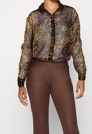 Przezroczysta bluzka na długi rękaw w zwierzęcy wzór, zestawiona z wysokim stanem i prążkowanymi brązowymi spodniami, modelka pokazana od szyi do połowy uda.