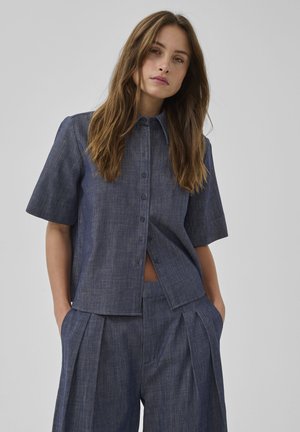 Femme aux longs cheveux bruns portant une chemise bleue à manches courtes boutonnée et un pantalon large plissé assorti, les mains dans les poches.