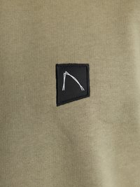 Tela de color verde oliva con una fina textura de canalé; presenta un parche rectangular negro con un diseño abstracto de líneas blancas.