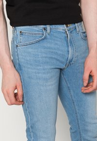 Jeans in denim azzurro chiaro con una vestibilità slim, caratterizzati da un design classico a cinque tasche e cuciture visibili. Indossati da una persona con una maglietta nera.