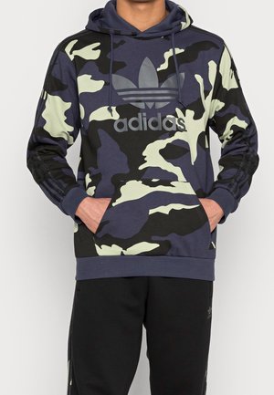 Uomo che indossa un hoodie Adidas a fantasia mimetica navy e nera con tasca a marsupio e logo Adidas sul petto, abbinato a pantaloni neri Adidas.
