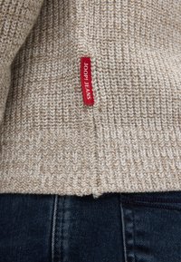 Beżowy sweter robiony na drutach z teksturowanym wzorem, z czerwoną metką "JOOP! JEANS" na dole, w połączeniu z ciemnoniebieskimi dżinsami.
