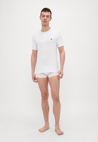 Balta trumpa rankovėmis henley marškinėliai su keturių mygtukų užtrauktuku, su mažyte juoda logotipu ant krūtinės; derinami su baltais boxer tipo apatiniais.