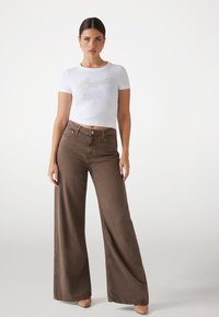 Top corto blanco con el logo "Guess" en plata, combinado con jeans marrones de cintura alta y pernera ancha. Tela suave y corte ajustado.