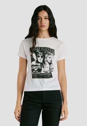Femme aux longs cheveux foncés portant un t-shirt blanc avec un motif graphique d'une photo en noir et blanc d'un groupe de musique, et un pantalon noir, sur un fond uni.