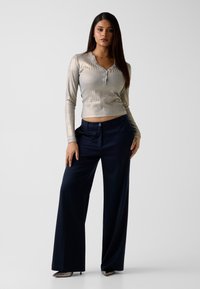 Metallic-goldener Langarm-Oberteil mit gerippter Textur und drei Knöpfen am Kragen, kombiniert mit dunkelblauen Hosen mit weitem Bein und silbernen High Heels.