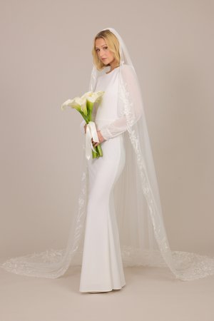 Braut in einem langärmeligen weißen Kleid und Schleier, die einen Strauß weißer Calla-Lilien vor einem schlichten beigen Hintergrund hält.