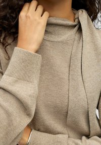 Gros plan sur une personne portant un pull en tricot beige avec un détail de cravate au cou, touchant le col d'une main.