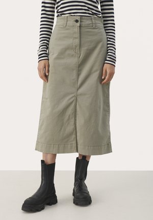 Femme portant une jupe midi beige avec boutons et fermeture éclair devant, des bottes noires épaisses et un haut rayé noir et blanc à manches longues.