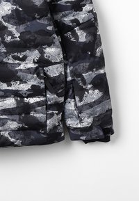 Přezkoušecí bunda v odstínech černé a šedé s texturovaným, abstraktním vzorem. Disponuje zipovou kapsou a žebrovanými manžetami pro teplo.