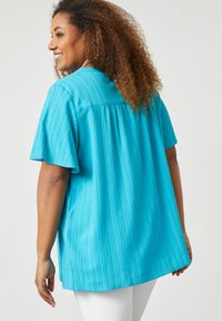 Blusa turchese a maniche corte con righe verticali, vestibilità comoda, orlo arrotondato e dettaglio plissettato sul retro. Indossata sopra pantaloni bianchi.