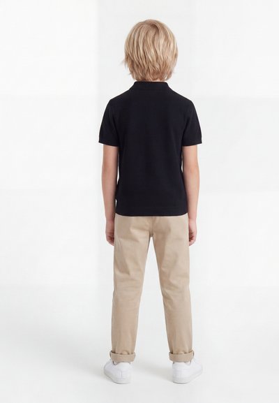 Ξανθό παιδί με πλάτη, φοράει μαύρο κοντομάνικο μπλουζάκι, μπεζ παντελόνια με γυρισμένα μπατζάκια και λευκά αθλητικά παπούτσια, στέκεται σε λευκό φόντο.