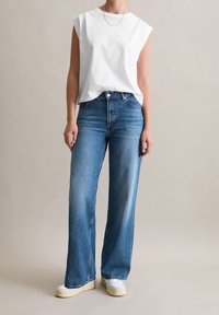 Top sin mangas de algodón blanco combinado con pantalones vaqueros de pierna ancha azul. Los vaqueros cuentan con cinco bolsillos y pasadores para cinturón; el calzado es unas zapatillas blancas.